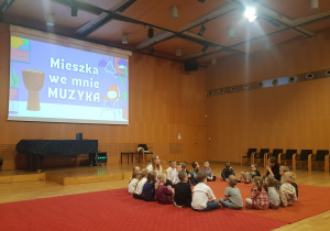 Grupa czerwona siedząca z animatorką w trakcie warsztatów ( mała sala w Filharmonii -czerwony dywan-fortepian )