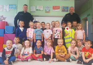 GRUPA CZERWONA- dzieci siedzą i stoją, pozując do wspólnego zdjęcia z policjantami
