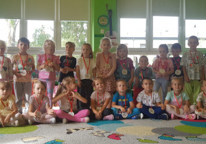 DZIECI Z GRUPY CZERWONEJ POZUJĄ DO ZDJĘCIA W SWOJEJ SALI Z ZAWIESZINYMI MEDALAMI PO UROCZYSTOŚCI DNIA PRZEDSZKOLAKA. wŚRÓD NICH ZNAJDUJE SIĘ CHŁOPIEC Z DYPLOMEM ŚWIEŻO PASOWANY NA PRZEDSZKOLAKA
