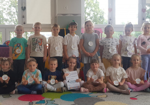 DZIECI Z GRUPY ŻÓŁTEJ POZUJĄ DO ZDJĘCIA W SWOJEJ SALI Z ZAWIESZINYMI MEDALAMI PO UROCZYSTOŚCI DNIA PRZEDSZKOLAKA. wŚRÓD NICH ZNAJDUJE SIĘ CHŁOPIEC Z DYPLOMEM ŚWIEŻO PASOWANY NA PRZEDSZKOLAKA